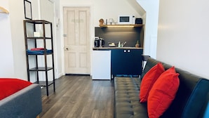 Un espacio habitable pequeño con kitchenette, un sofá con cojines rojos, piso de madera y un estante pequeño.