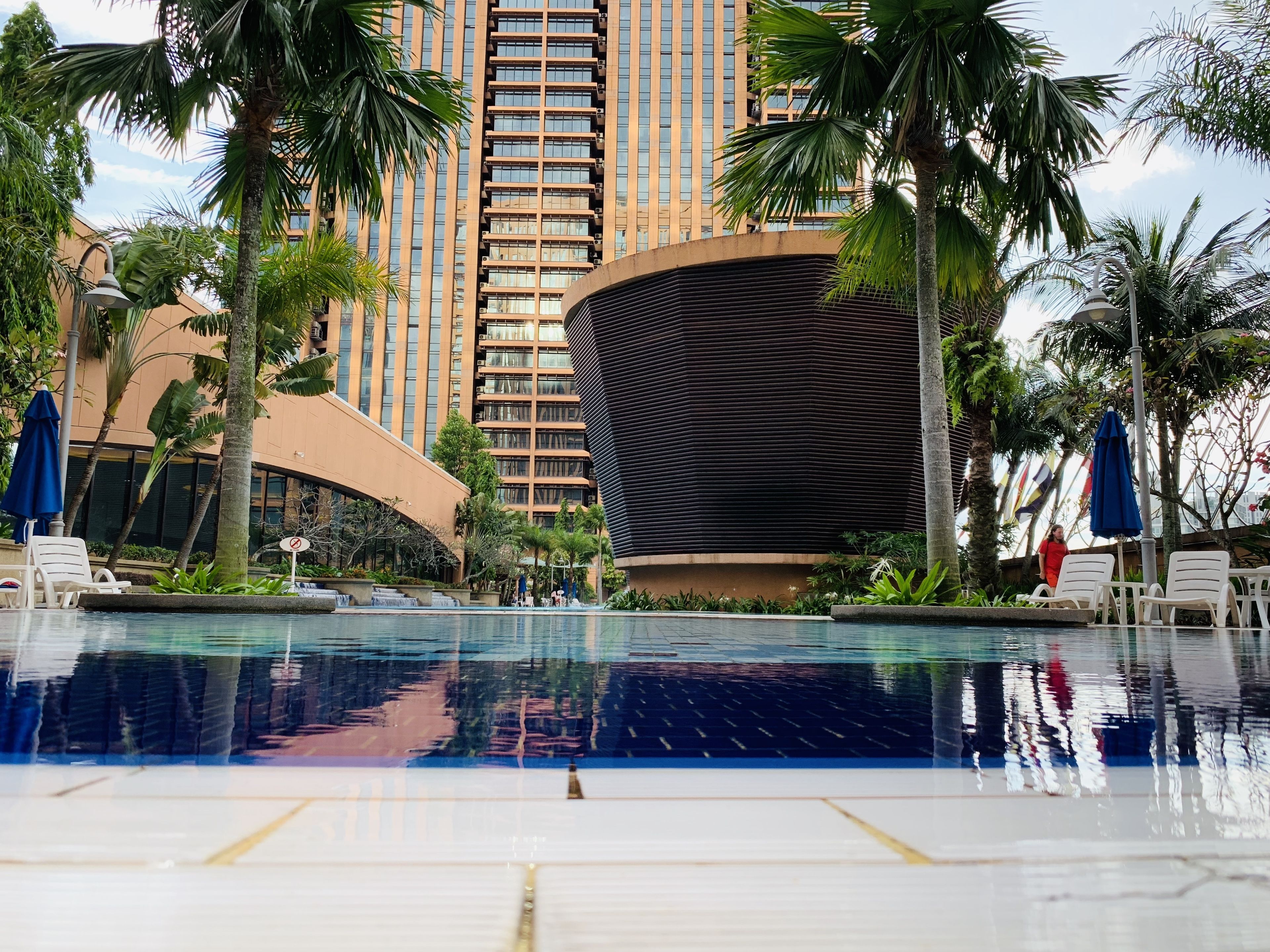 A modern hotel pool area with a large pool, palm trees, and a tall building in the background.