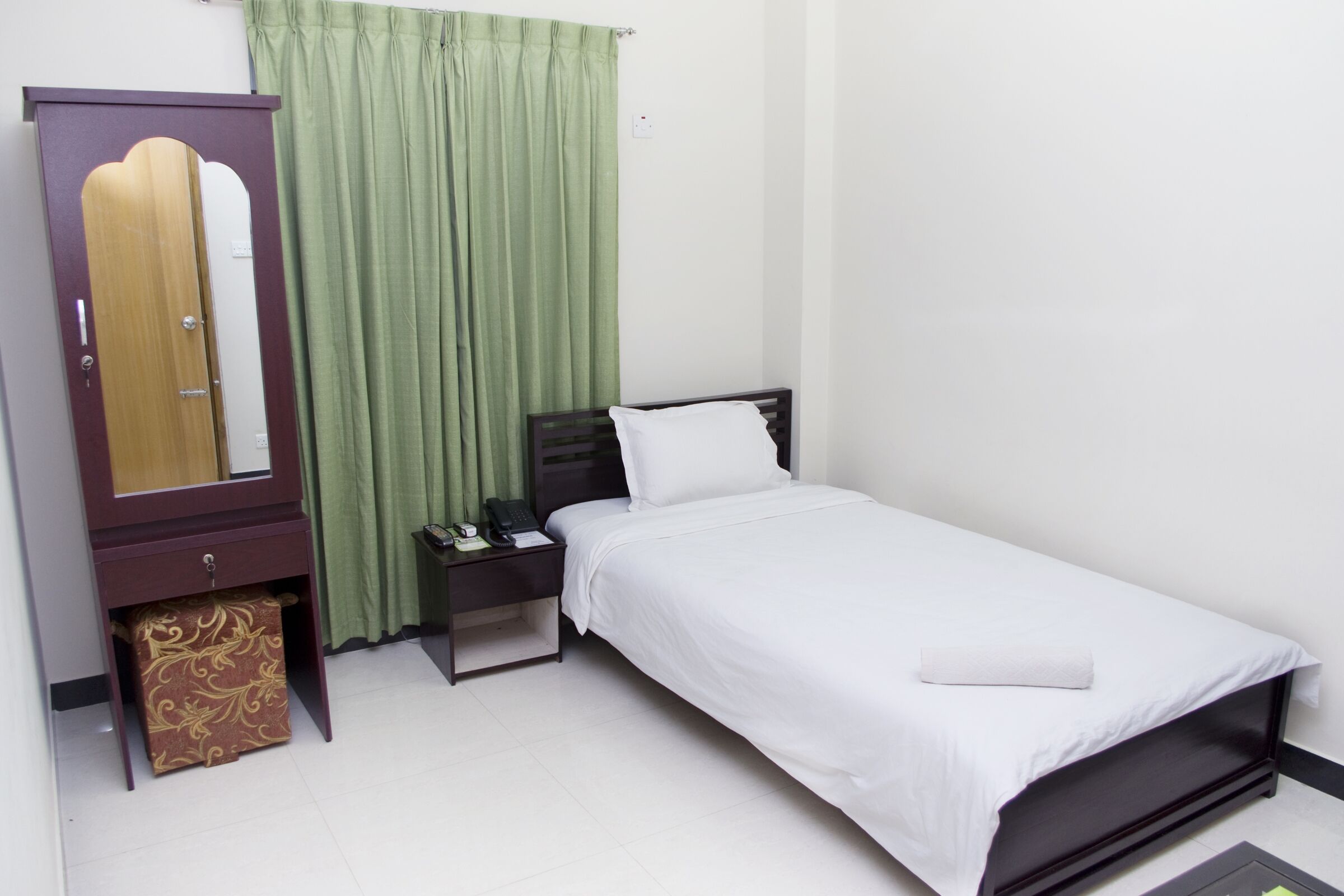 A single bed with a white mattress and pillow, a wooden wardrobe with a mirror, a green curtain, and a small bedside table with a telephone and papers.