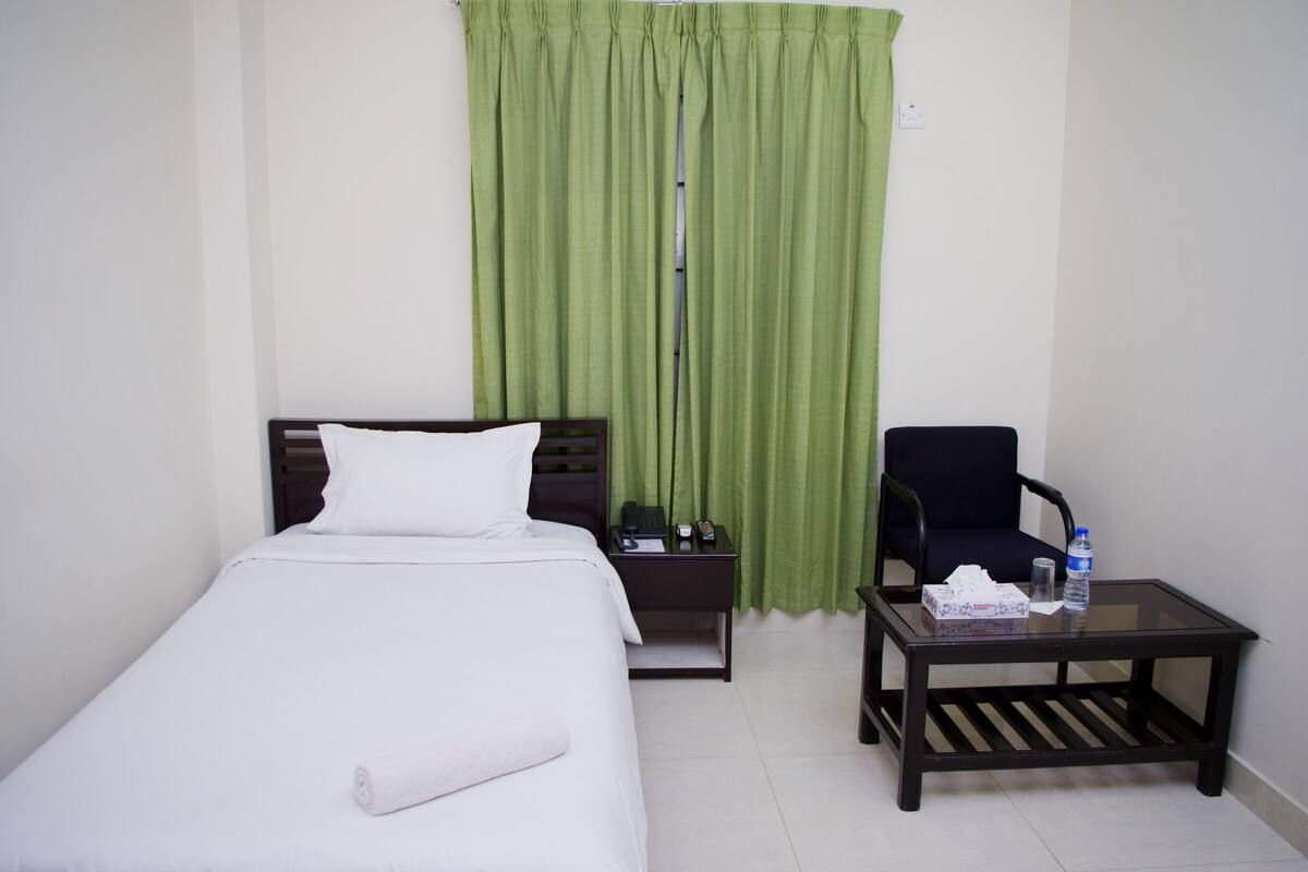 Une chambre d’hôtel avec un lit simple, une table de chevet avec un téléphone et une lampe, une chaise et une petite table sur laquelle se trouvent une bouteille et des mouchoirs.