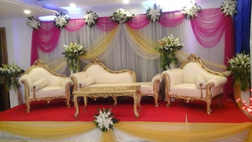 Um palco com dois sofás elaborados e uma mesa, coberta com tecido branco e rosa, decorada com arranjos florais.