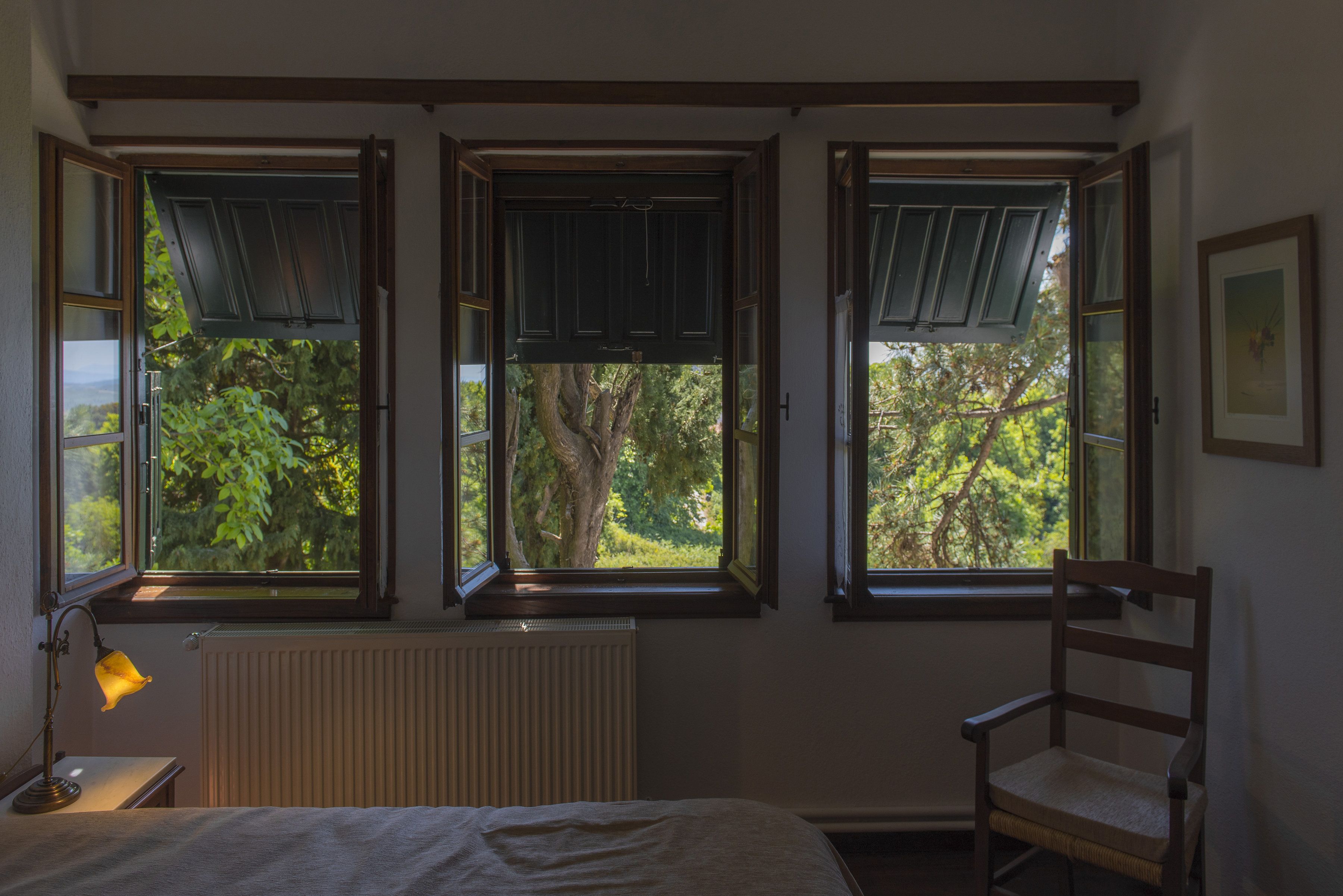 Une chambre avec trois fenêtres, un radiateur, une table de chevet avec une lampe et une chaise.