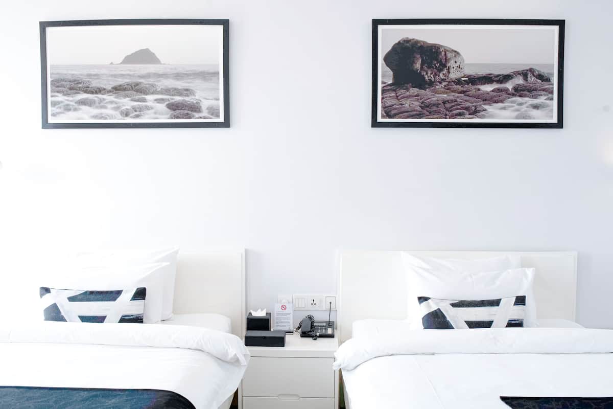 A hotel room with two beds, white bedding, a nightstand with a phone and a tissue box, and two framed pictures of coastal scenes on the wall.