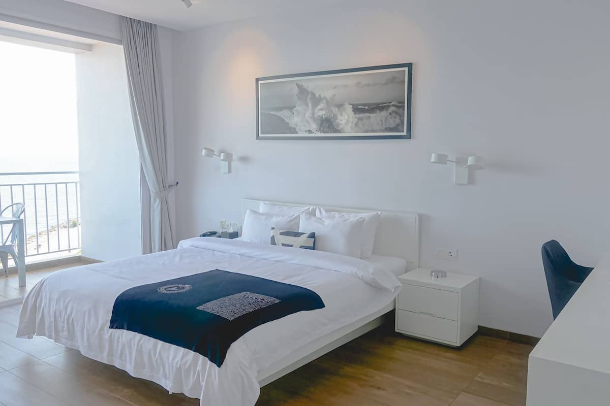 A hotel room with a bed, bedside tables, a desk, a chair, a window with curtains, and a wall-mounted painting.