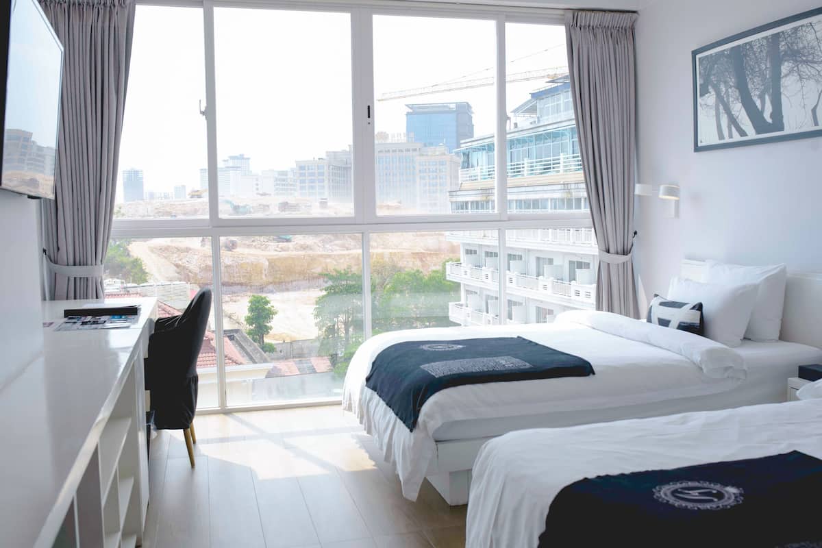 A hotel room with two beds, a desk, and a large window offering a view of the cityscape and construction site.