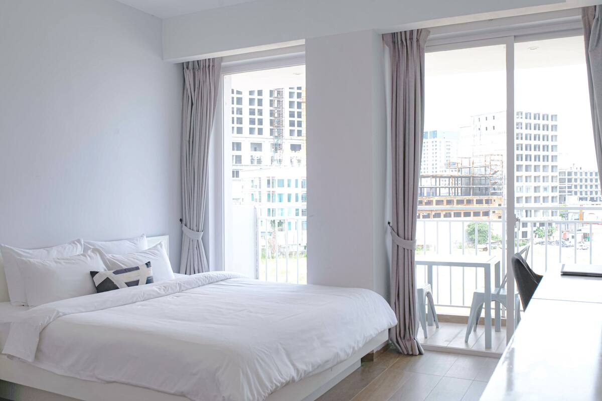 A modern hotel room with a large bed, a balcony with a view of the city, and a dining area with a table and chairs.