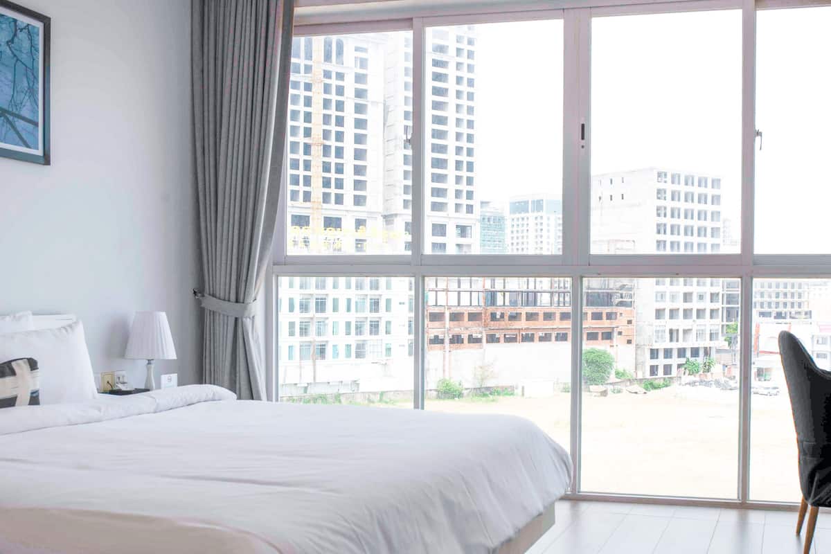 A hotel room with a large bed, a nightstand, a lamp, and a view of tall buildings through large windows.