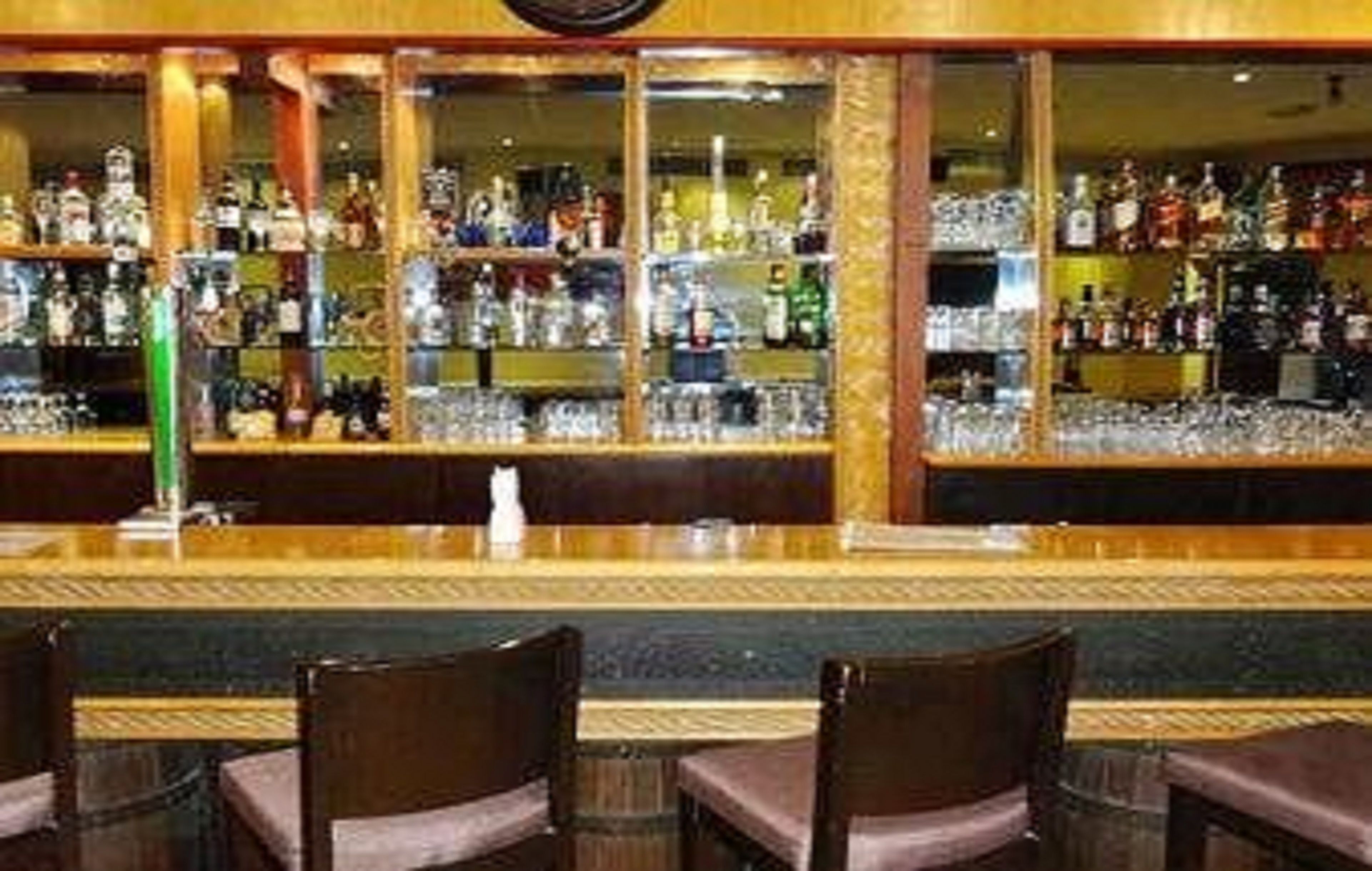 Een bar met flessen drank en glazen, houten stoelen en een toog.