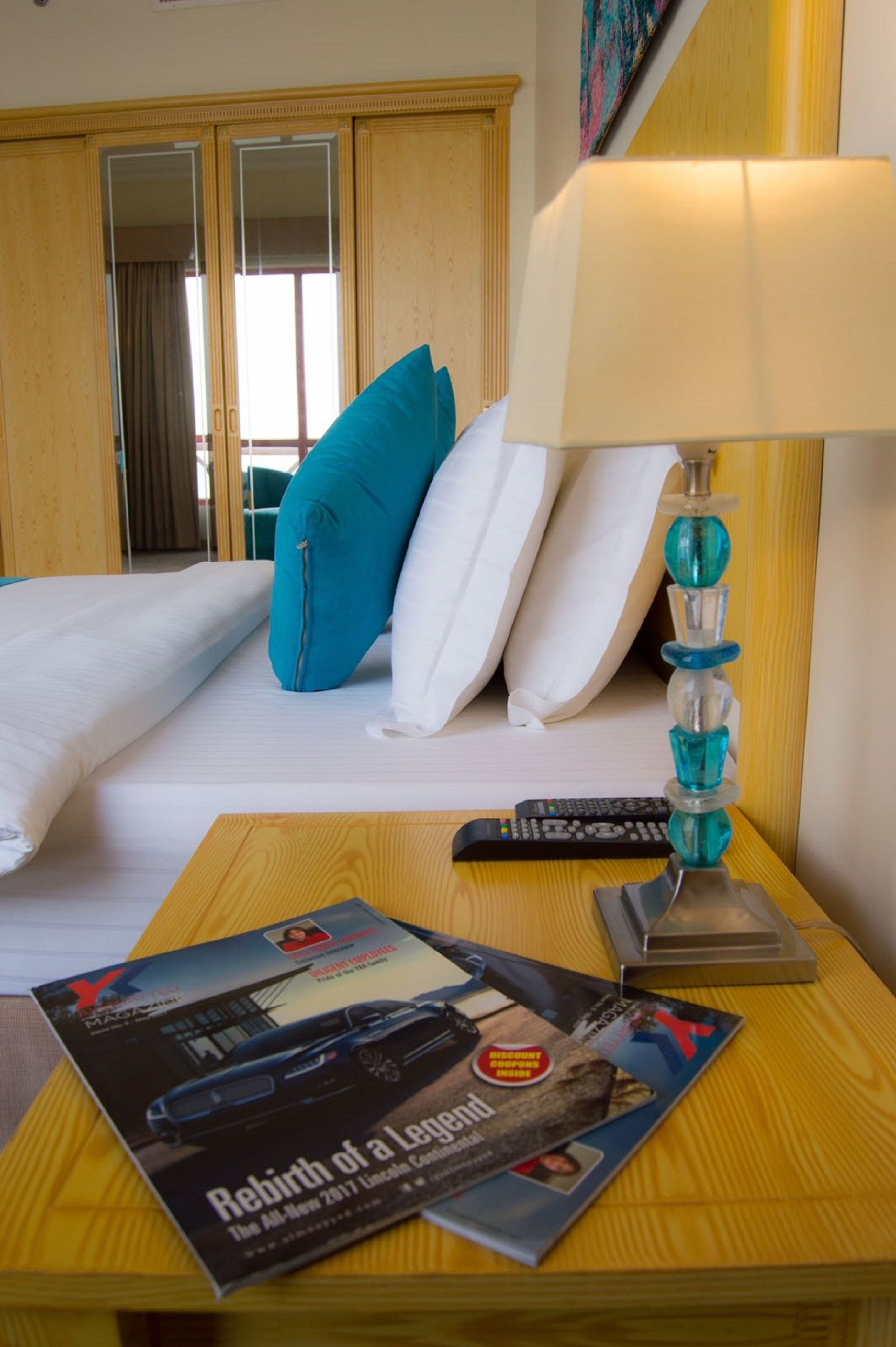 A hotel room with a bed, a wooden nightstand, a lamp, and a magazine on the nightstand.
