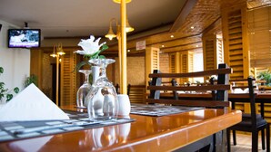 Interior de restaurante con mobiliario de madera, un televisor montado en la pared y una mesa dispuesta con servilleta blanca, cristalería y un arreglo floral.
