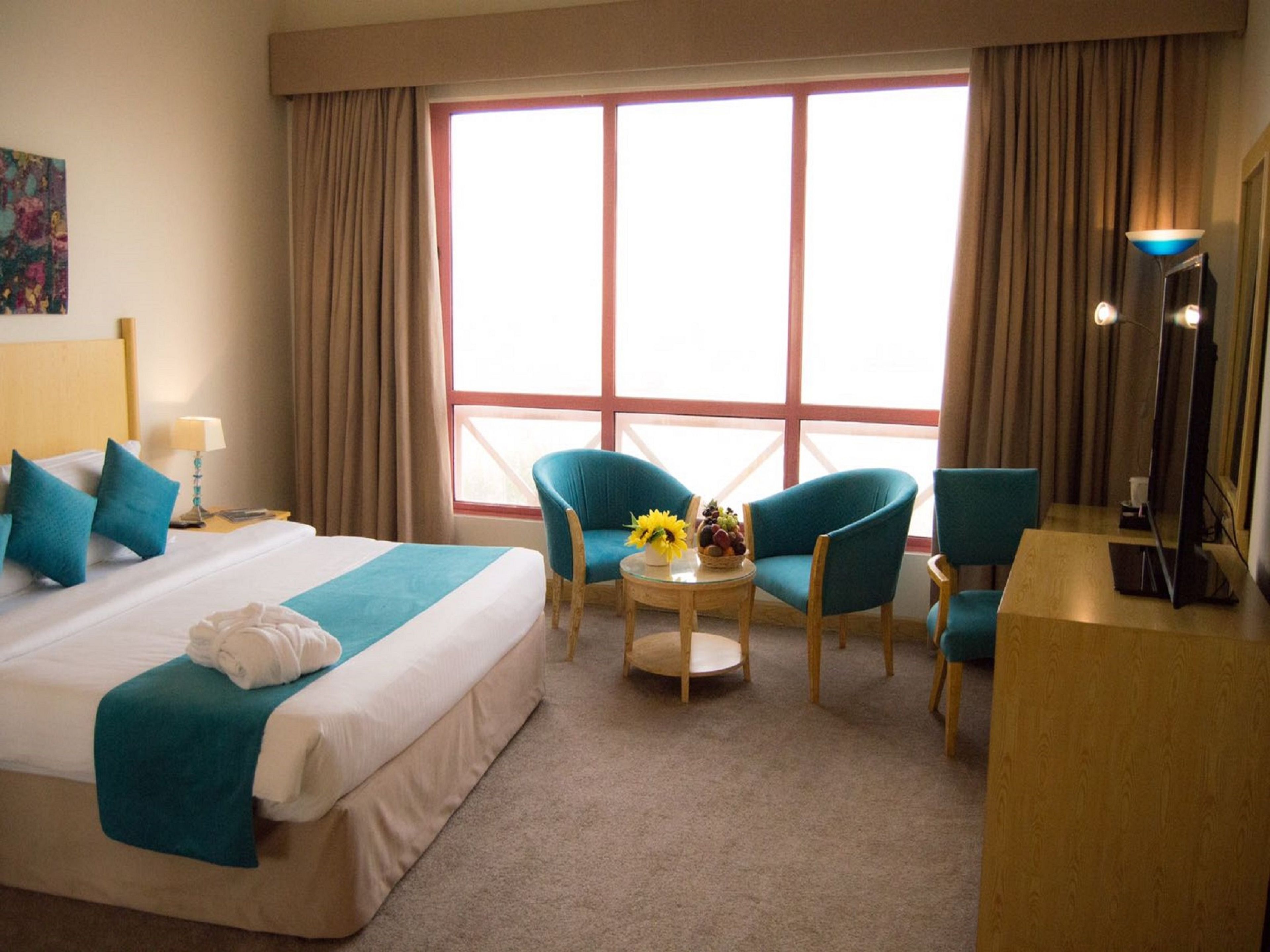 A hotel room with a large bed, two blue armchairs, a small round table with a fruit bowl, and a desk with a lamp.