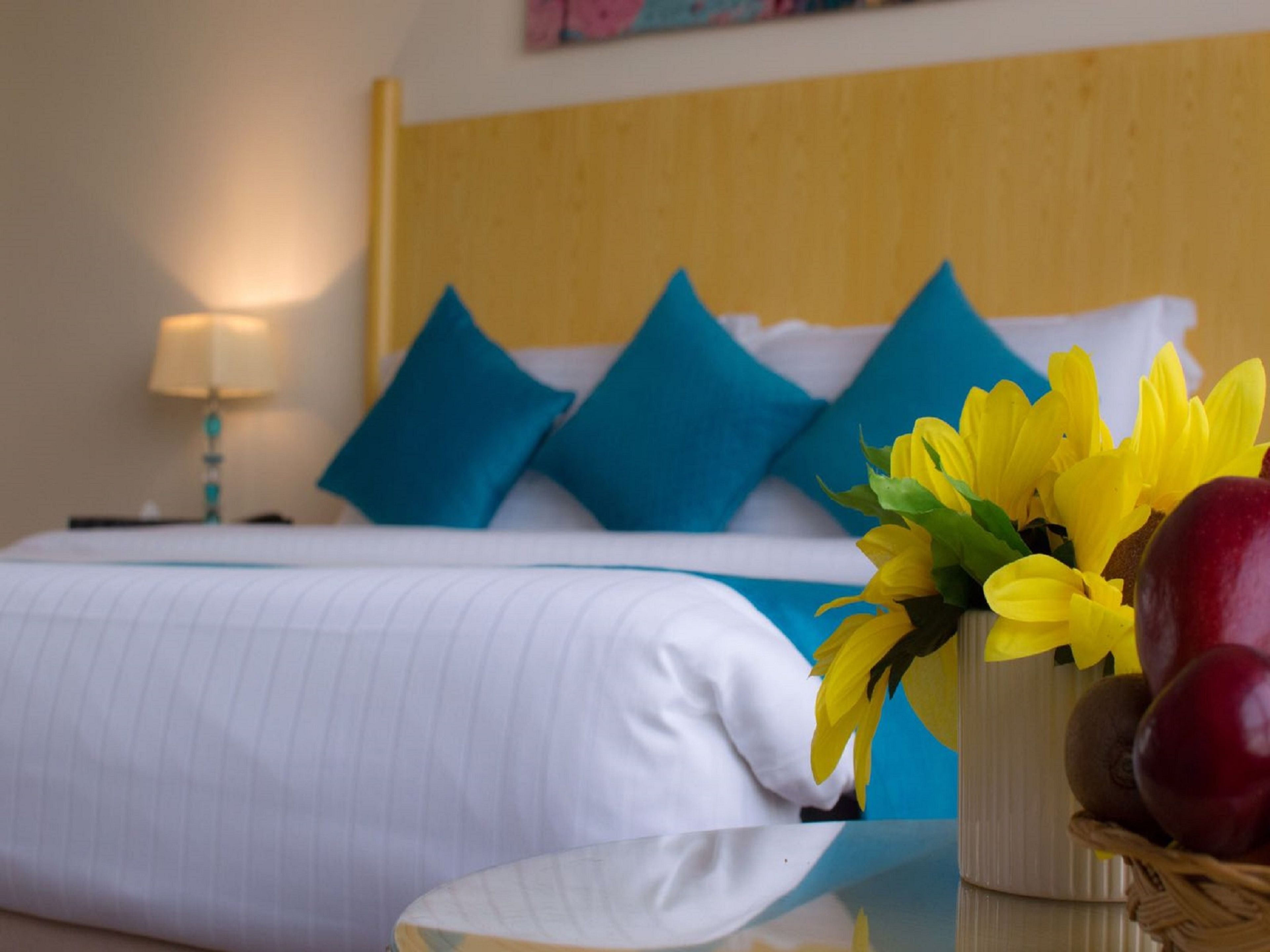 A hotel room with a bed, pillows, a bedside table, and a vase with flowers and fruits.