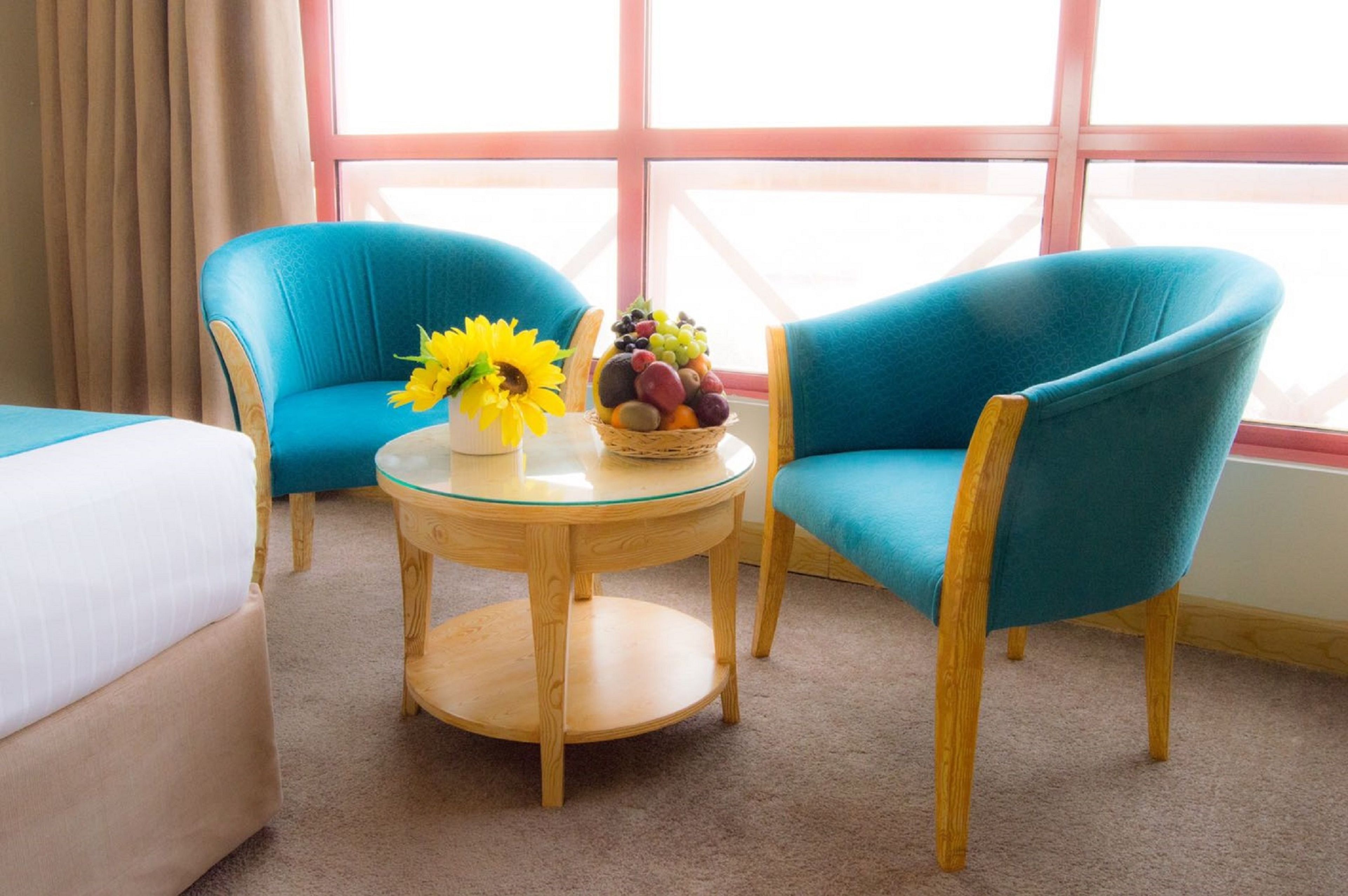 A room with two teal armchairs, a round wooden table with a glass top, a small vase with sunflowers, and a fruit bowl.