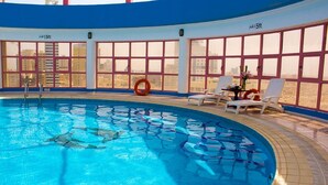 Una piscina interior con agua cristalina y azul, rodeada de grandes ventanales que ofrecen vistas de la ciudad.