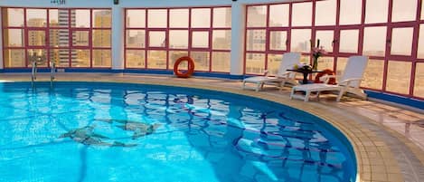 Una piscina interior con agua cristalina y azul, rodeada de grandes ventanales que ofrecen vistas de la ciudad.