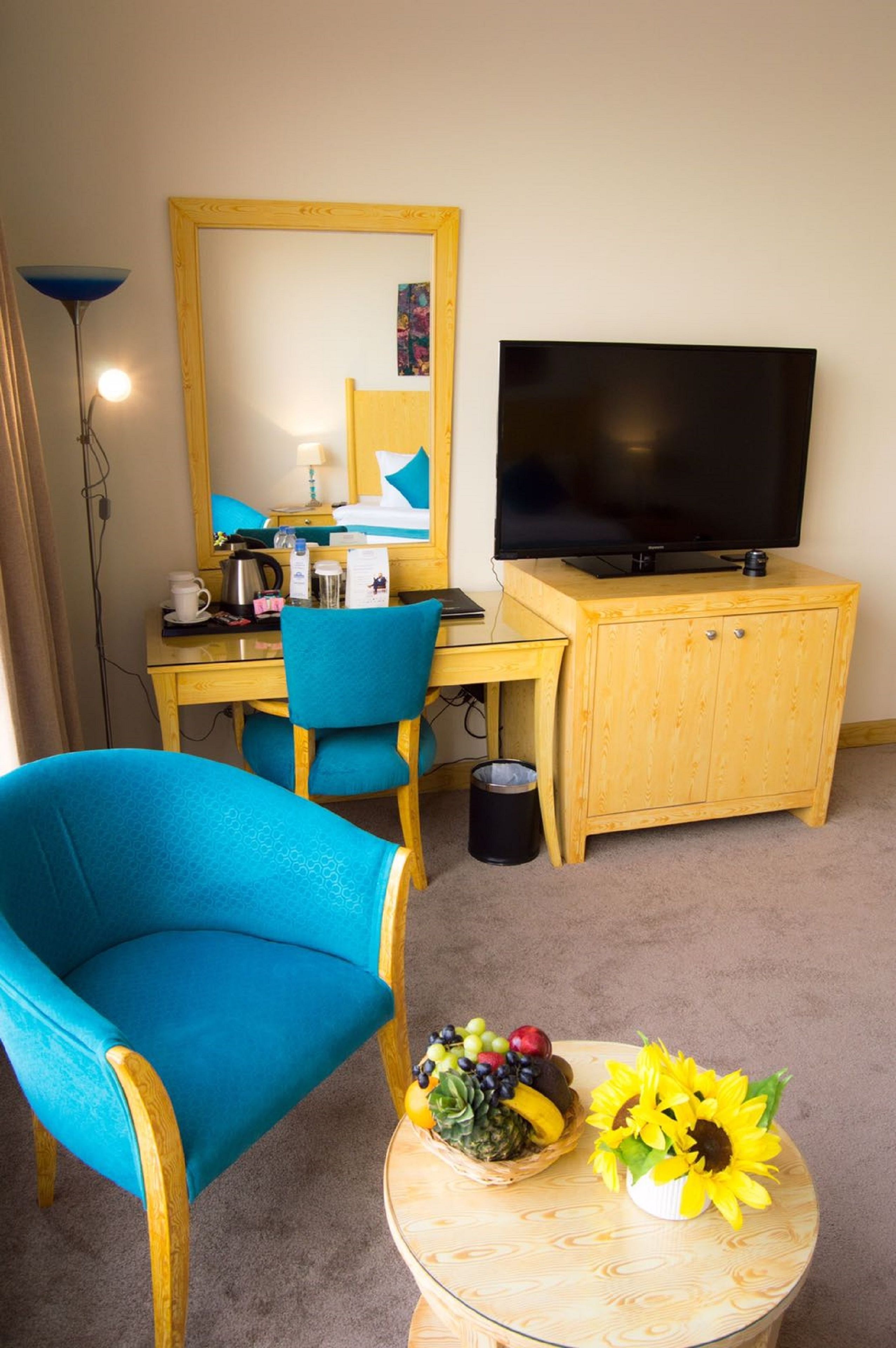Une chambre d’hôtel équipée d’un bureau, d’une chaise, d’une télévision, d’une petite armoire, d’une poubelle et d’une table ronde sur laquelle se trouvent un panier de fruits et un tournesol.