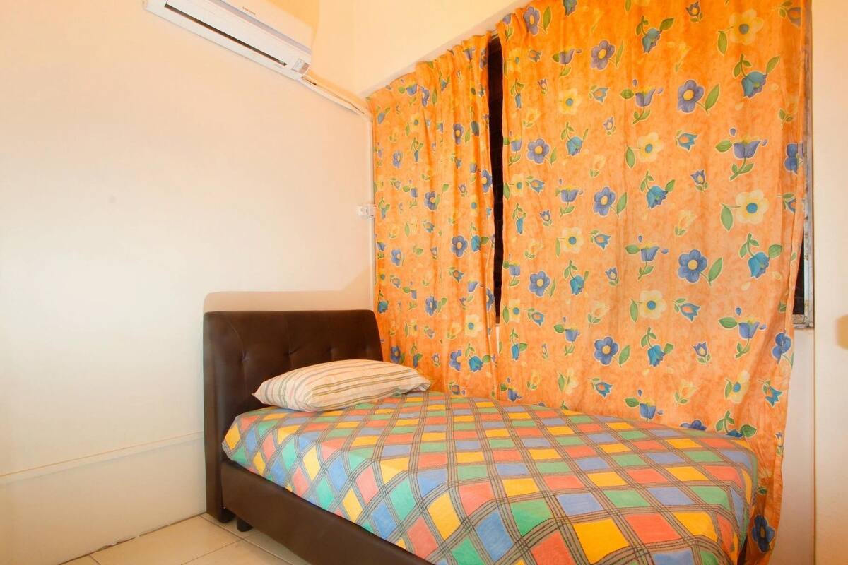 Ein kleines Zimmer mit einem Einzelbett, einem braunen Kopfteil, einer farbenfrohen karierten Bettdecke und einem orangefarbenen Blumenvorhang.