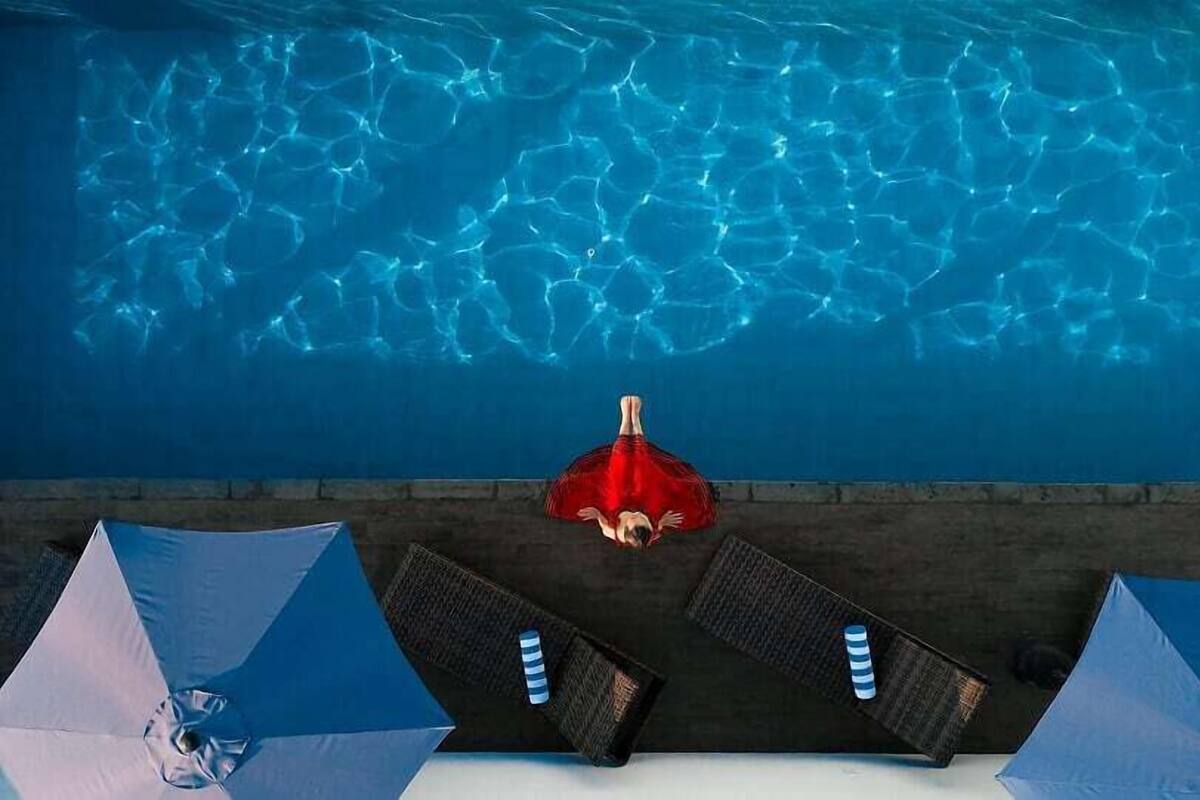 Uma piscina com água cristalina, uma pessoa de fato de banho vermelho e cadeiras de praia com guarda-sóis.
