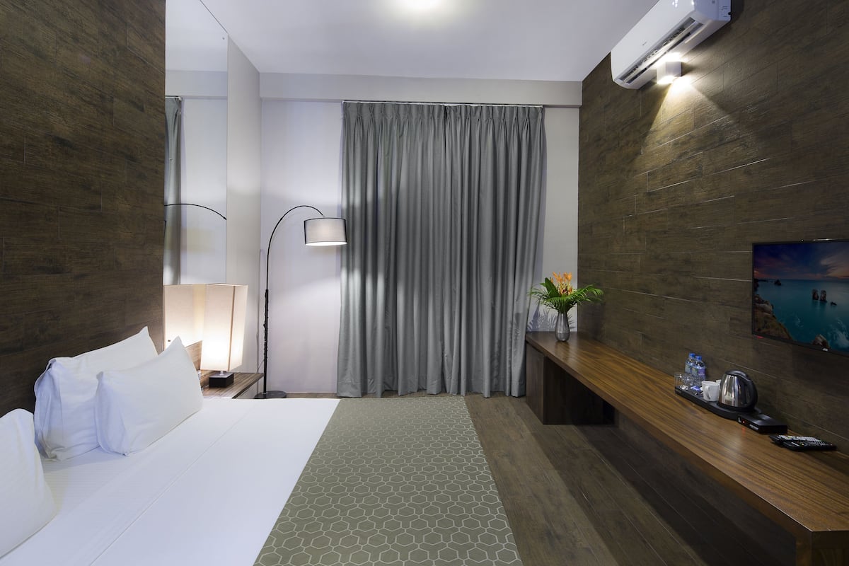 Quarto de hotel com uma cama, uma escrivaninha com luminária, uma televisão e um ar condicionado.