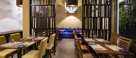 Un restaurante con mesas y sillas de madera, un banco acolchado azul y un amplio ventanal con vistas a zonas verdes.