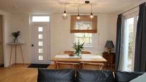 A dining area with a wooden table, wicker chairs, and a vase with plants. There is a door with a window above it, and a lamp on a side table.