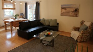 A modern living room with a glass coffee table, black sofa, and floral armchair. There is a dining area with a table and chairs, and a painting on the wall.