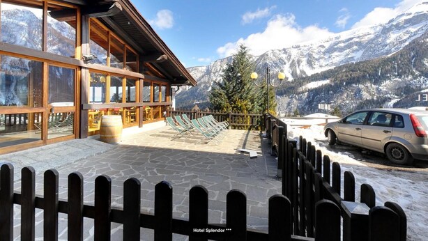 A hotel with a wooden exterior, large windows, a stone patio with lounge chairs, and a view of snow-capped mountains. - Hotel Splendor (Dimaro Folgarida)