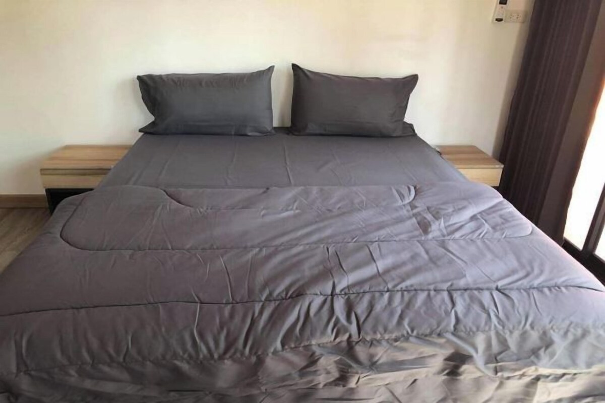 Un lit avec une couette grise et deux oreillers, une table de chevet en bois à gauche et une fenêtre avec des rideaux à droite.
