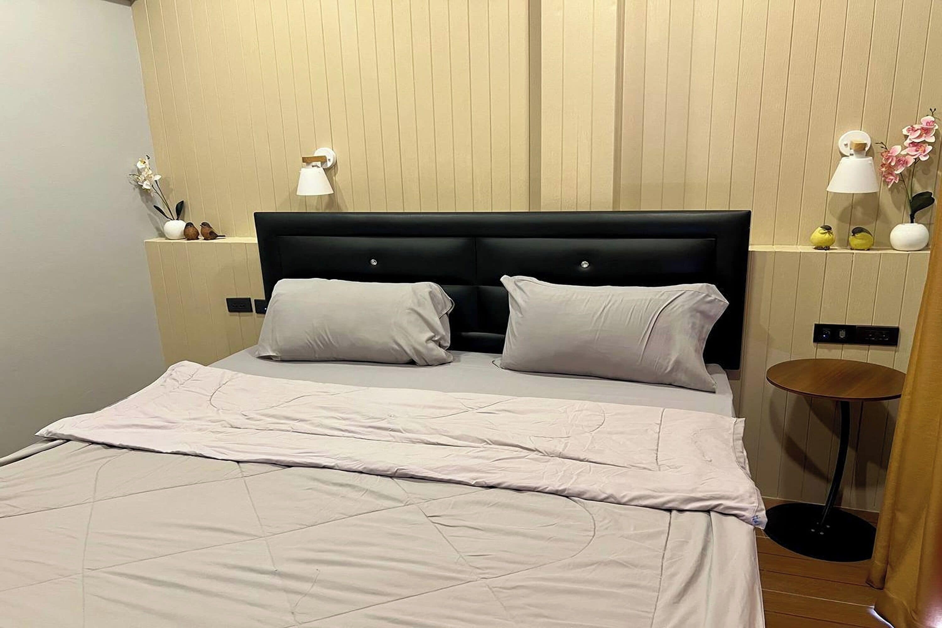 Una cama bien hecha con cabecera negra, dos mesitas de noche con lámparas y una pequeña mesa redonda con una silla.