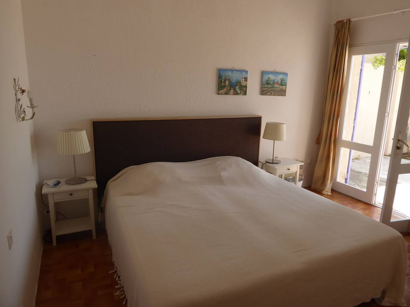 Een slaapkamer met een groot bed, twee nachtkastjes met lampen, een deur met gordijn en twee aan de muur bevestigde schilderijen.