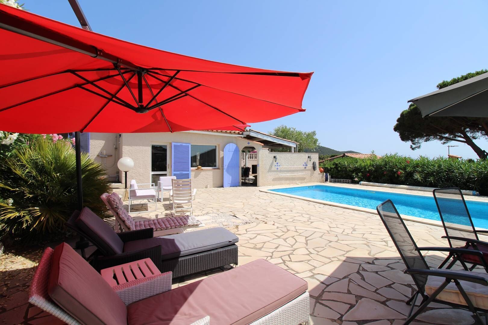 Un espace aménagé au bord de la piscine, avec des chaises longues, un parasol rouge et un ciel d’un bleu limpide.