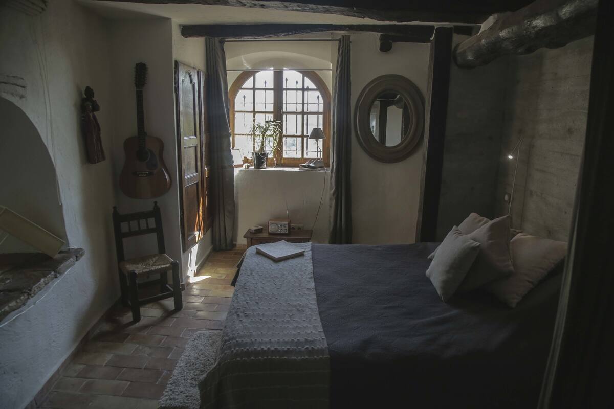 A bedroom with a bed, a guitar, a chair, a window with curtains, and a small table with a lamp.