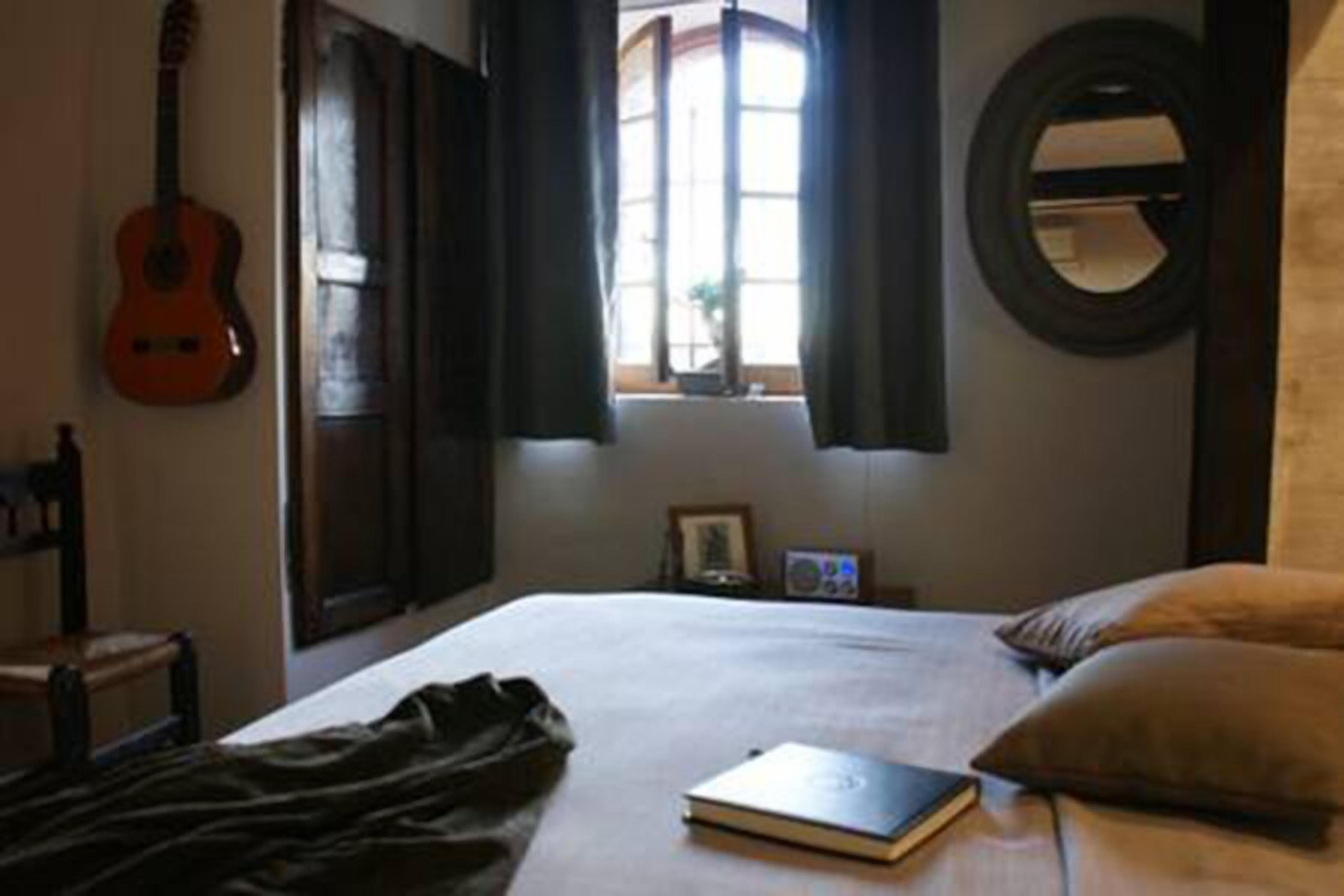 Une chambre avec un lit, une guitare, un miroir rond et une fenêtre avec des rideaux.