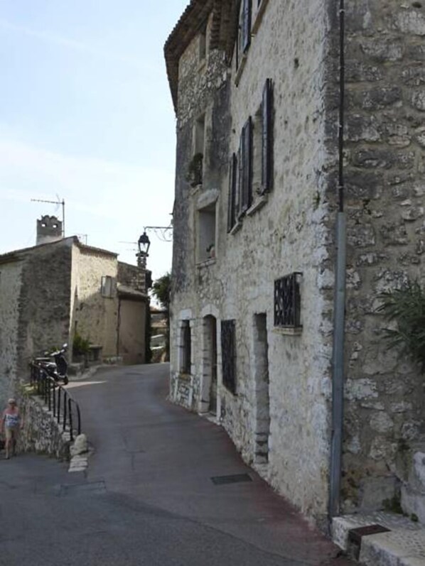 A narrow street with stone buildings, a metal fence, and a person standing by the entrance. - La Maison Aux Bonsaïs (Saint-Paul-de-Vence)