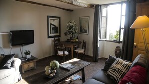 Uma sala de estar com sofá, mesa de centro, televisão, mesa de jantar e cadeiras, e uma janela com cortinas.