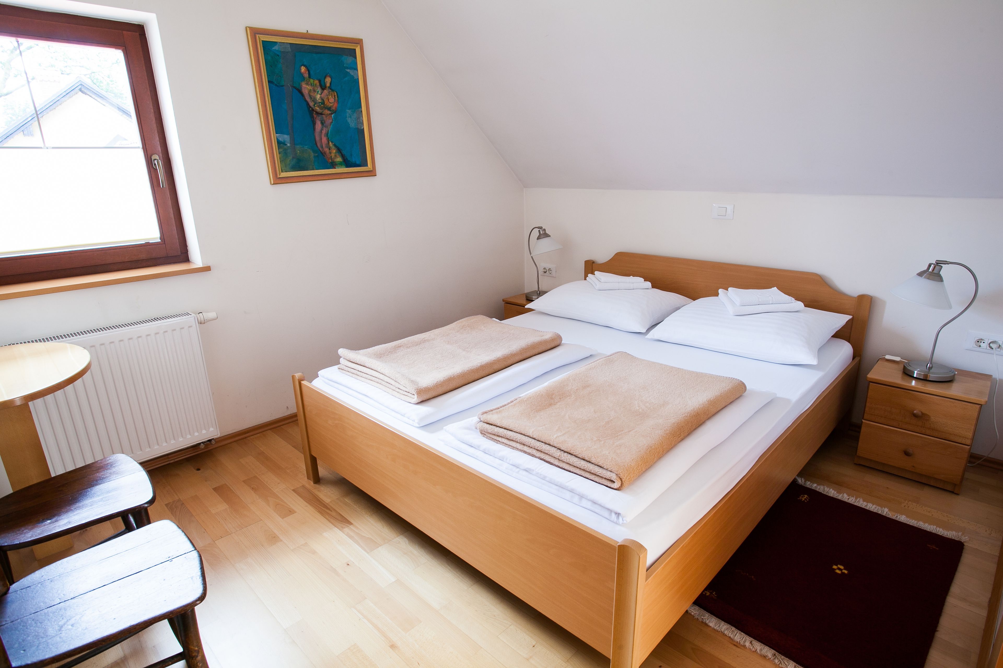 A neatly made bed with two folded towels, a wooden nightstand with a lamp, a small table, and a radiator.