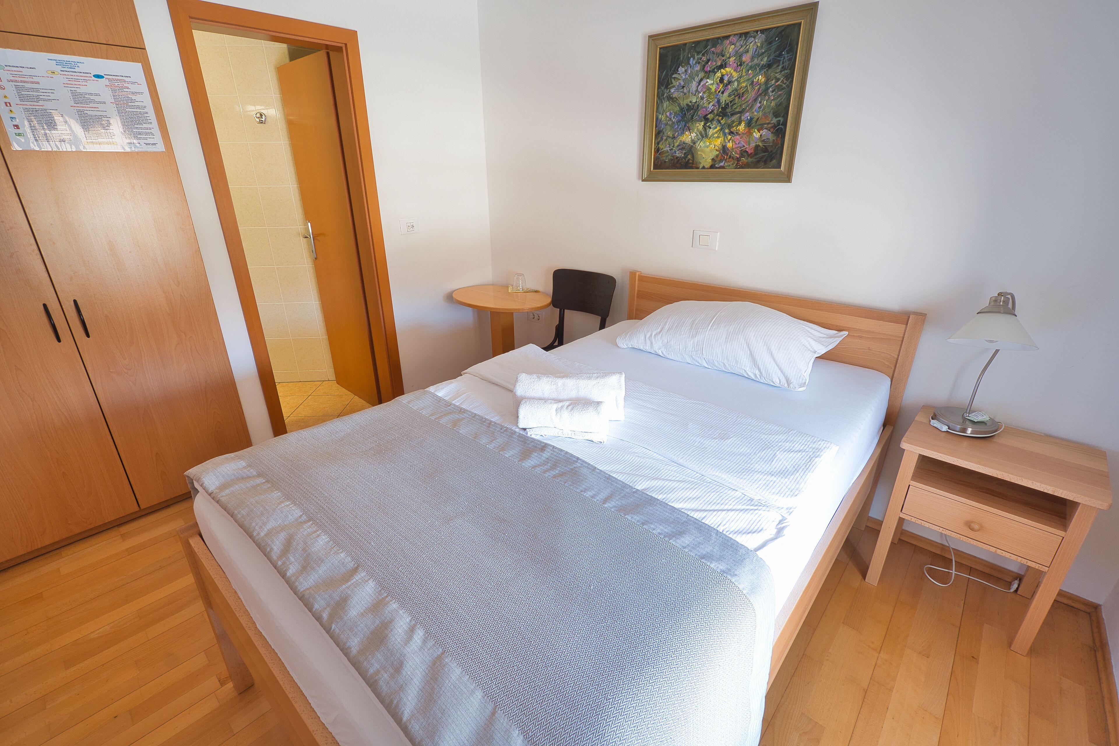 A neatly made bed with a white comforter and pillows, a wooden nightstand with a lamp, a small round table with a glass, and a door leading to another room.