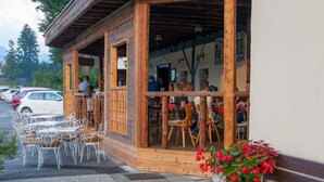 Ein Café im Freien mit Holztischen und -stühlen, einem überdachten Sitzbereich und Blicken auf parkende Autos und Bäume im Hintergrund.