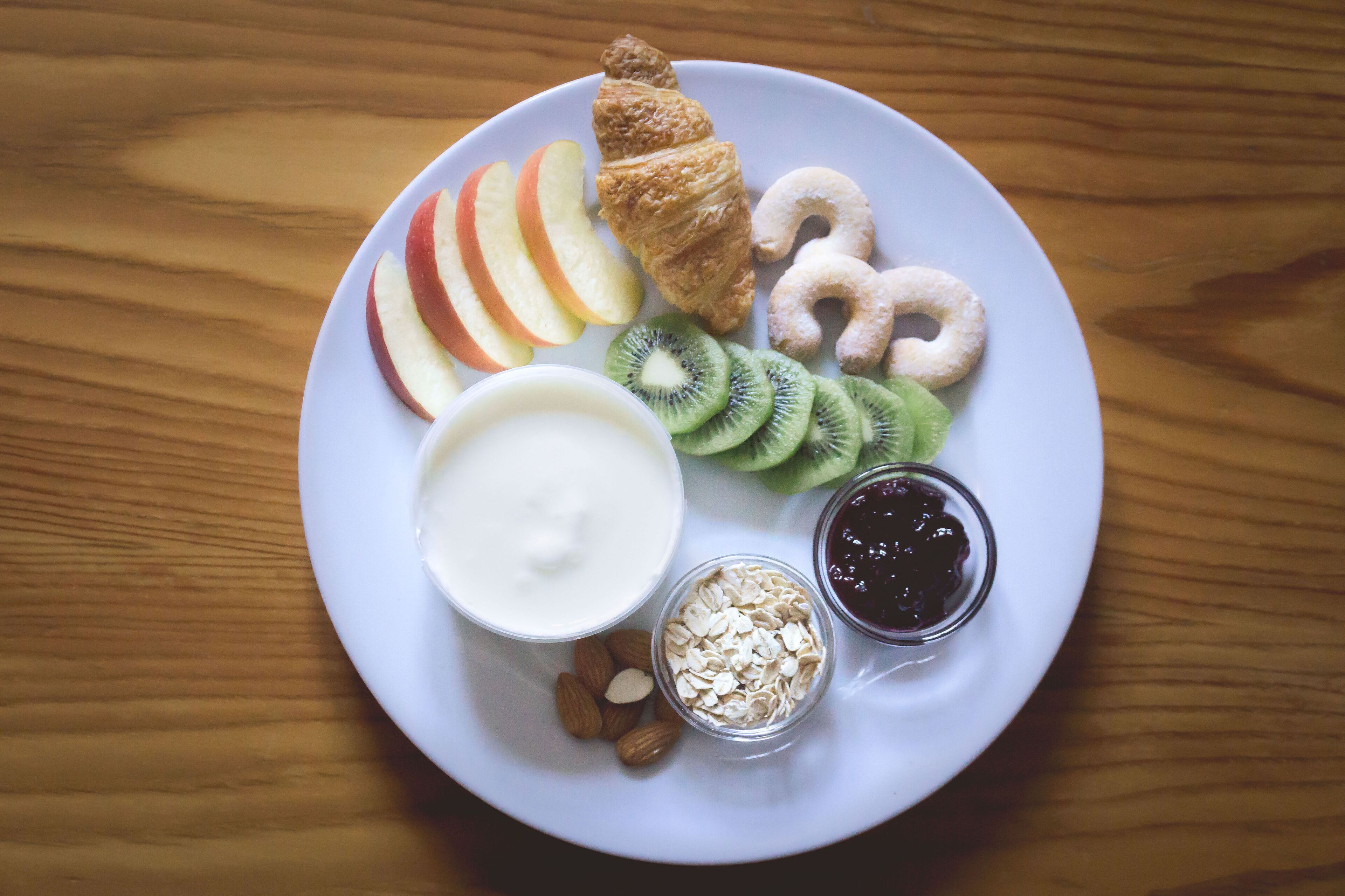 Un plato con rodajas de manzana, kiwi, almendras, un croissant, yogur y galletas sobre una mesa de madera.