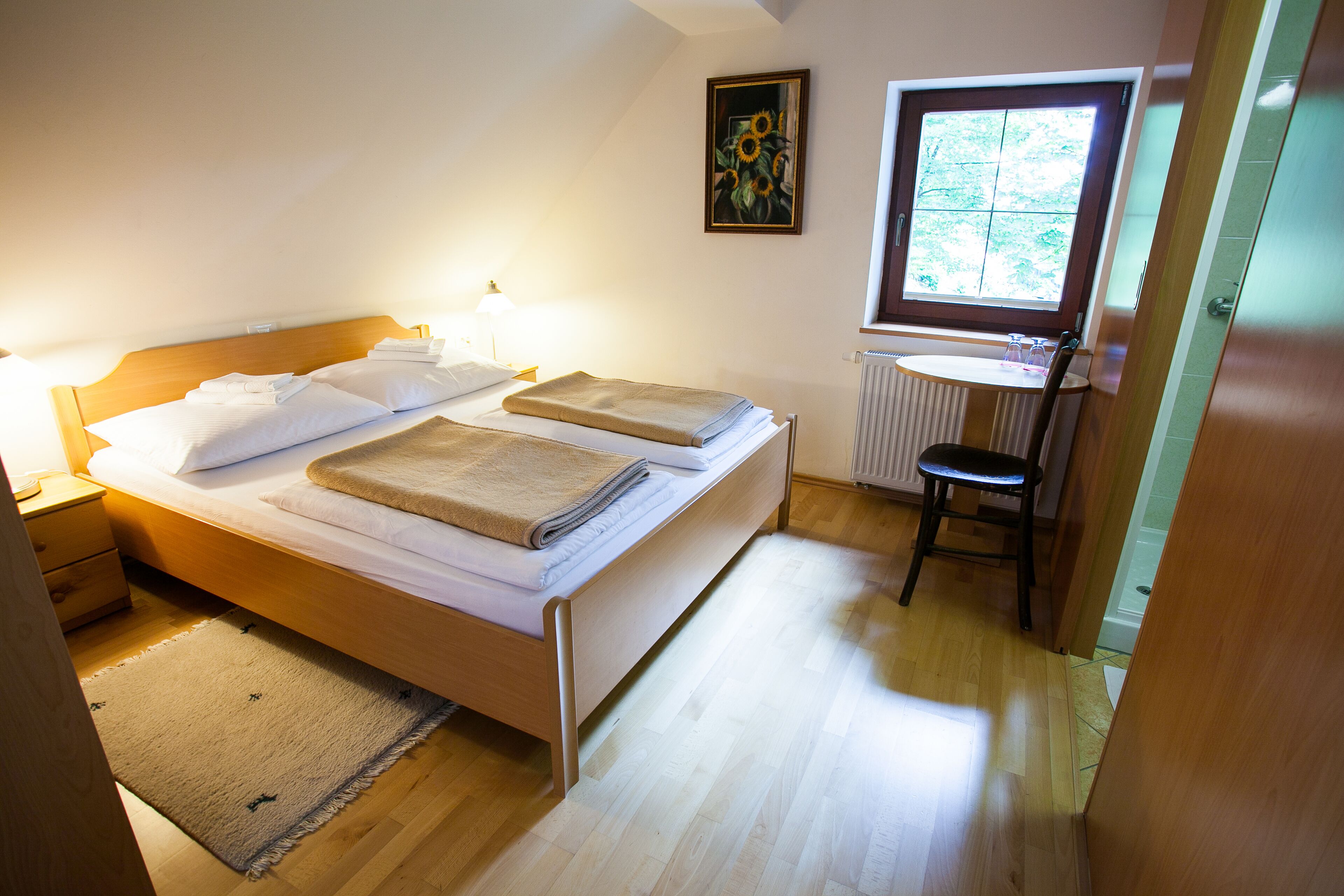 A double bed with a wooden headboard, a bedside table with a lamp, a small stool, a radiator, and a window with a view of greenery.