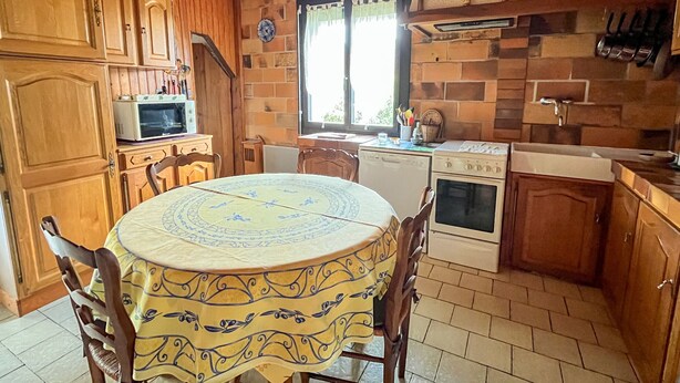 Une cuisine avec des meubles en bois, une table à manger recouverte d’une nappe à motifs, un four à micro-ondes, une cuisinière et une fenêtre avec des rideaux.