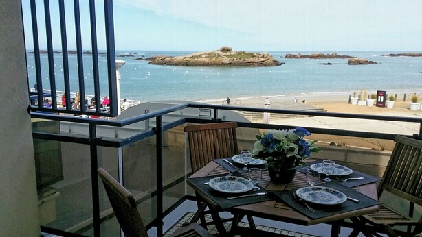 A balcony with a table set for two, overlooking a beach with rocks and the sea.