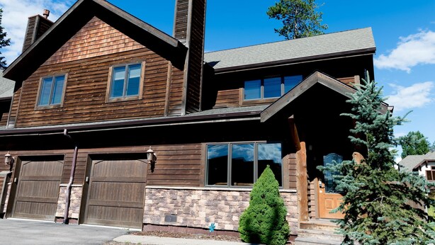 A two-story wooden house with a chimney, large garage doors, and a well-maintained lawn.