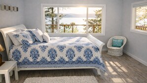 Una camera da letto con un'ampia finestra che si affaccia su un lago e montagne, un letto con lenzuola azzurre a motivi decorativi, una sedia in vimini con cuscino e un tappeto bianco.
