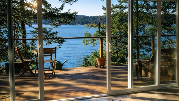 Un ampio balcone con vista su un lago, pavimento in legno e mobili da esterno tra cui sedie e un tavolo.