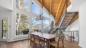 Un'ampia sala da pranzo con un grande tavolo in legno e sedie, un lampadario e vista su alberi attraverso ampie finestre.
