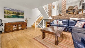 Ein geräumiges Wohnzimmer mit Holzboden, einem großen Gemälde einer Berglandschaft, einem Kamin aus Ziegelsteinen und einem Ledersofa.