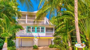 A two-story house with a balcony, surrounded by palm trees and a paved driveway.