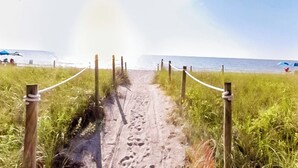 A sandy beach path with footprints leading to the ocean, surrounded by tall grass and wooden posts with ropes.