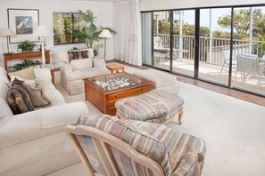 Un salon spacieux avec un grand canapé, une table basse et des fauteuils à rayures. Il y a un balcon avec des meubles d’extérieur et une vue sur le paysage.